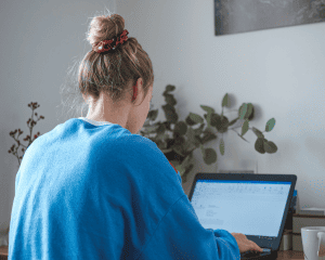 young female wearing a blue jumper, sitting at a desk, looking at a laptop