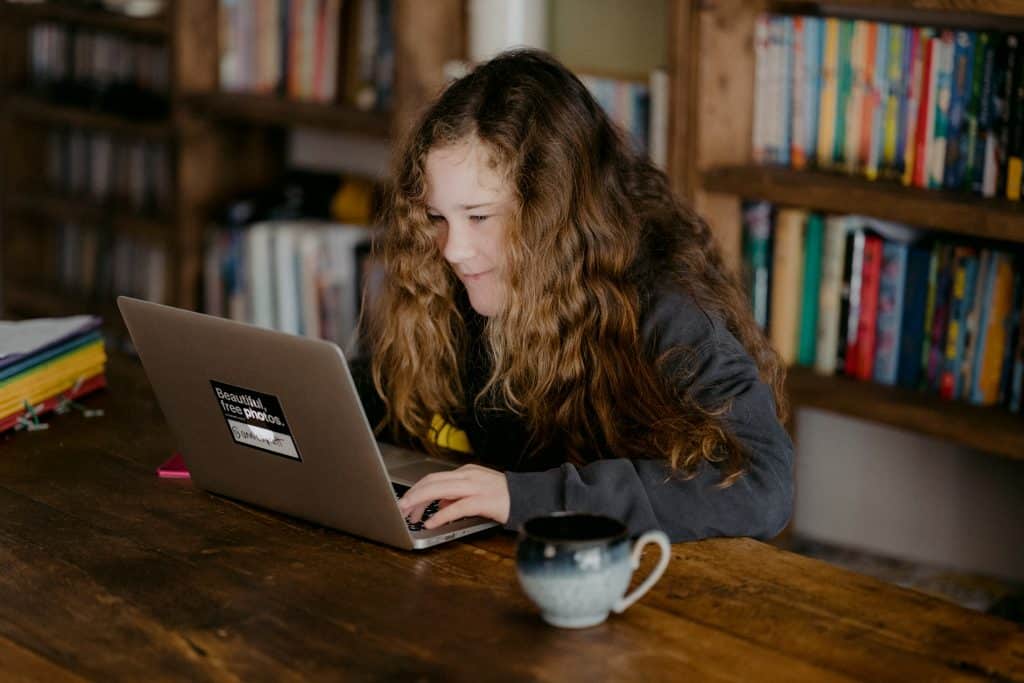 Young female child working on a laptop with a bookcase behind her.