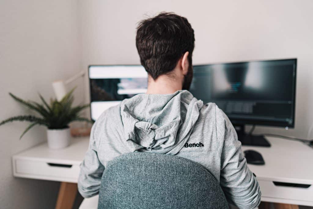 young male with beard and grey hoodie sitting at a desk looking at a desktop.