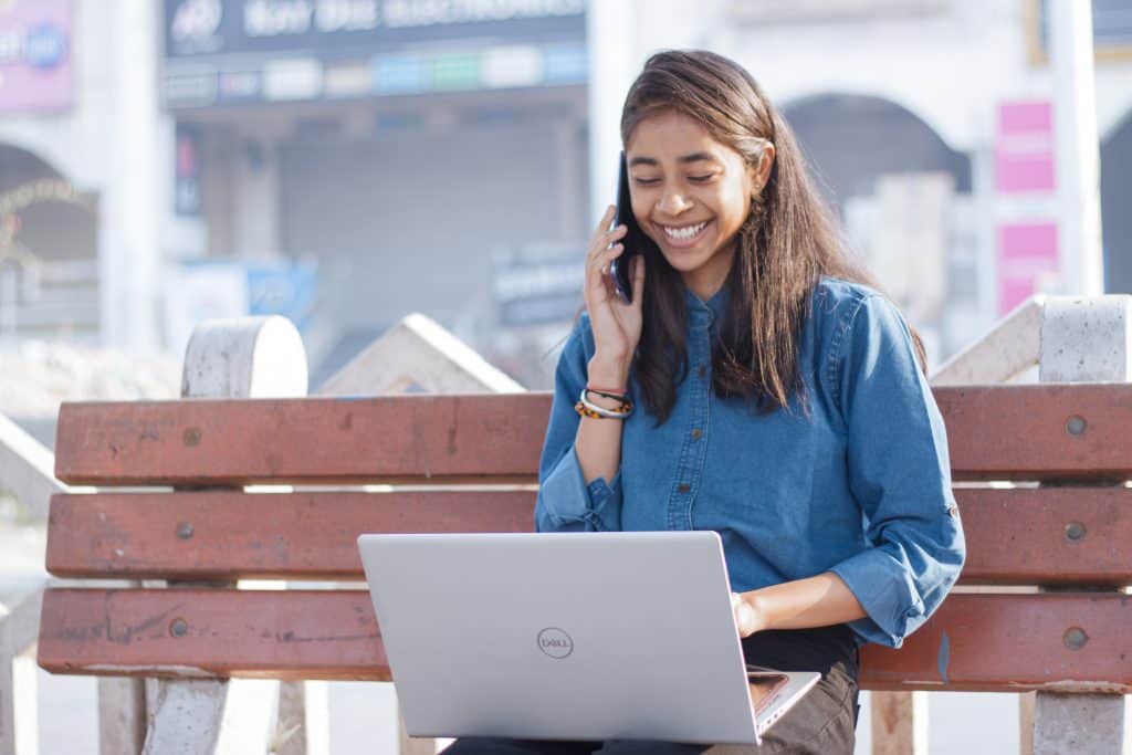 Smiling woman on bench, on phone and on a laptop