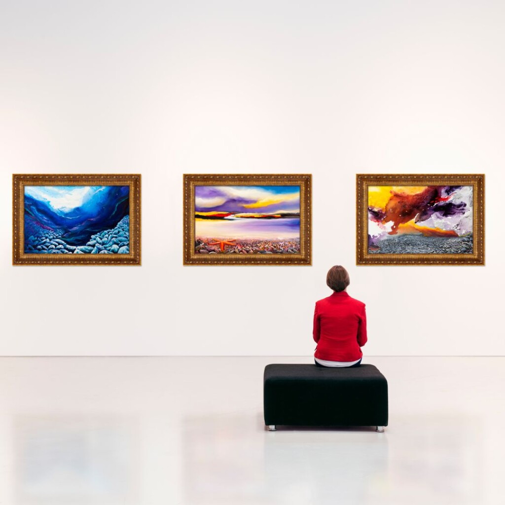 Online Art History Course with NEC. Female in red jack sat on black stool admiring a row of paintings in an art gallery