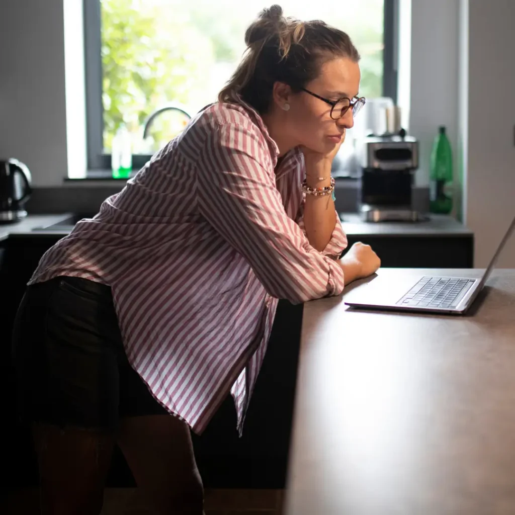 Woman leans on kitchen counter top and studies her laptop
