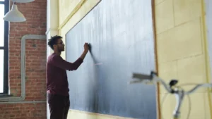 A man writing on a large chalkboard in a classroom, representing the first steps in how to become a teacher and the qualifications needed for a career in education.