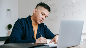 A tutor sits at a desk engaged with a laptop, taking notes with a pen while working in a bright, organised space. A whiteboard with maths equations is in the background, reflecting an academic setting.