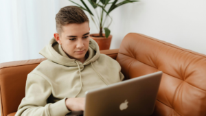Teenage student in a hoodie studying on a laptop from a sofa, representing relaxed, flexible learning with distance education and AS level courses.