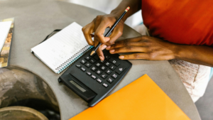 Close-up of hands using a calculator next to a notepad and orange folder, symbolising practical maths use in education, finances, and career planning.