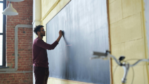 A man writing on a large chalkboard in a classroom, representing the first steps in becoming a teacher and the qualifications needed for a career in education.