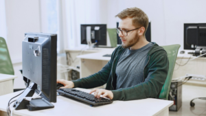 A man sits at a computer desk in a classroom environment, working on a desktop PC—representing careers and education in computing and computer science.