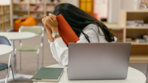 A student sits at a library table with her head down on a red book in frustration, illustrating the emotional impact of receiving disappointing GCSE results.