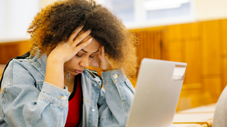 A student sits in a classroom with her head in her hands in front of a laptop, showing visible signs of stress during exam revision.