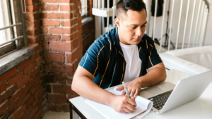 A focused adult student sitting at a desk in a bright room, writing notes in a notebook while looking at an open laptop—symbolising flexible learning and mature study.