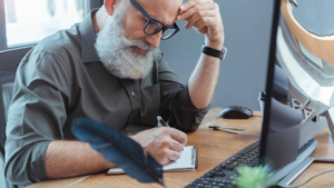 An older man with grey hair and glasses sitting at a desk, focused as he writes in a notebook beside a desktop computer—symbolising lifelong learning and academic determination.