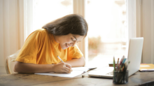 A smiling young woman studying at a desk near a window, writing in a notebook with a laptop and pencil pot in front of her—illustrating a positive and focused distance learning experience.