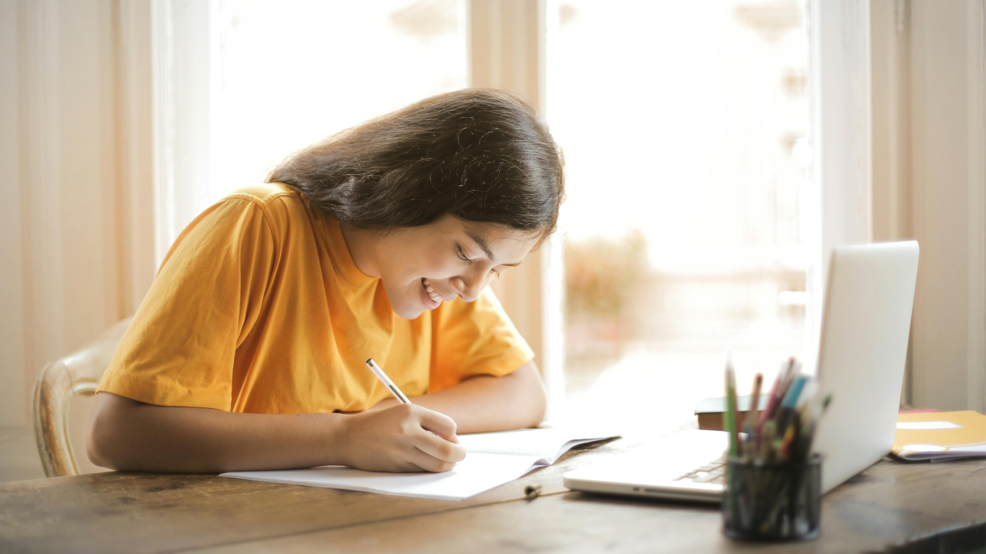 A smiling young woman studying at a desk near a window, writing in a notebook with a laptop and pencil pot in front of her—illustrating a positive and focused distance learning experience.