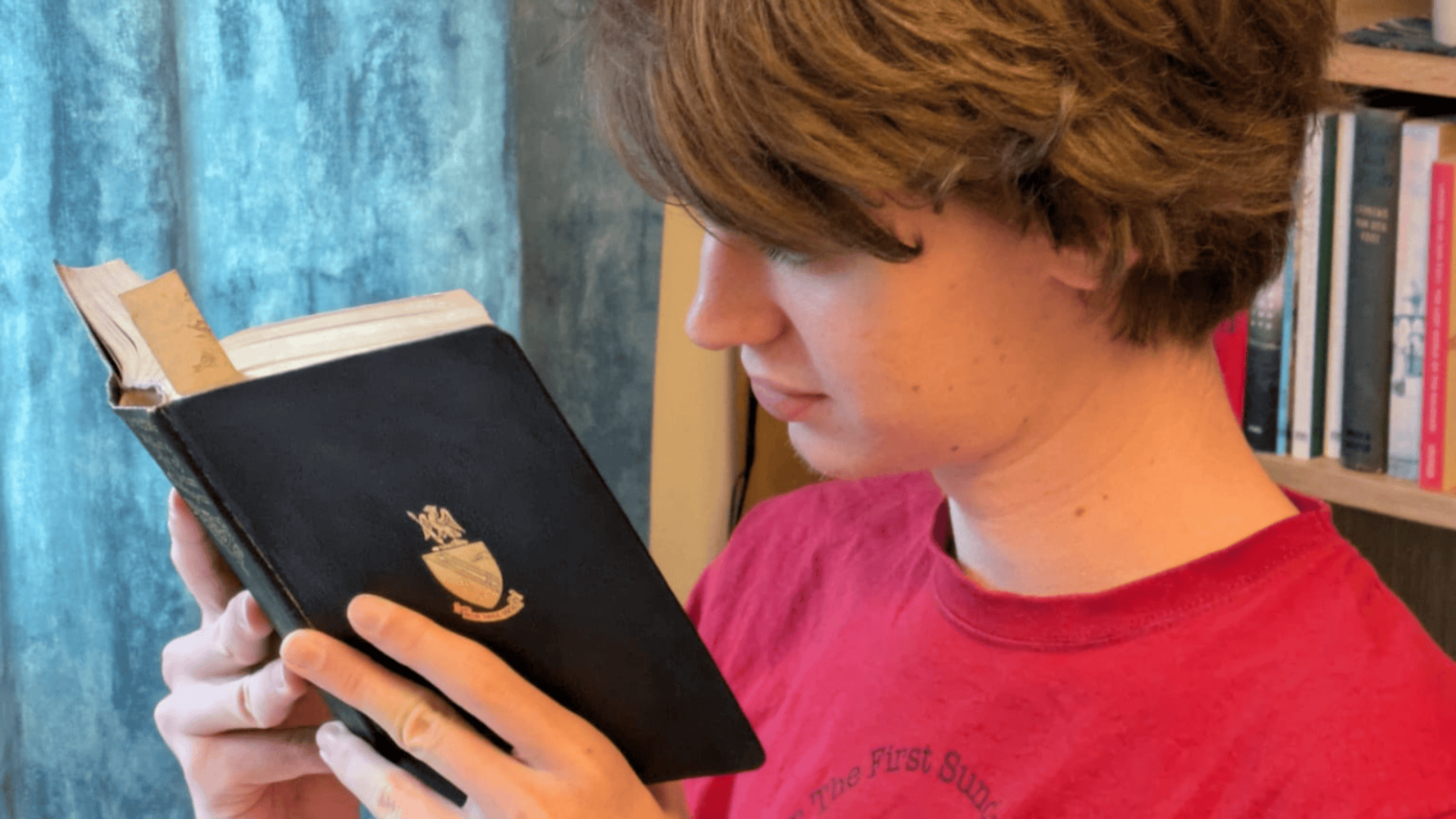 A young student in a red T-shirt is deeply focused on reading a black hardback book with a crest on the front. He stands in front of a blue curtain, beside a bookshelf filled with books. The scene reflects concentration, curiosity, and a love for literature.