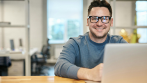 A smiling man wearing glasses sits at a desk using a laptop in a bright, modern workspace—conveying a positive and professional distance learning environment.
