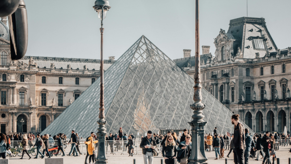 A busy day outside the Louvre Museum in Paris, with the iconic glass pyramid in view and people walking through the square—highlighting the cultural appeal of learning a new language for travel and connection.