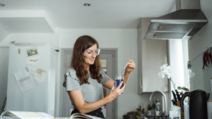A smiling woman wearing safety goggles performs a simple science experiment in her kitchen using a beaker and pipette—demonstrating how to study A level science from home