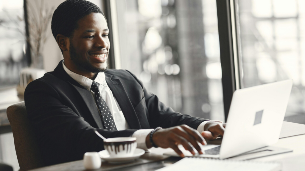 A smiling professional in a suit works on a laptop at a modern café, with a coffee beside him—reflecting career progression, flexibility, and success through distance learning CMI management qualification.