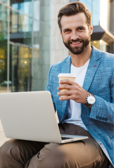 Tutor sits outside with his laptop on his knee and a coffee in his hand, smiing to the camera