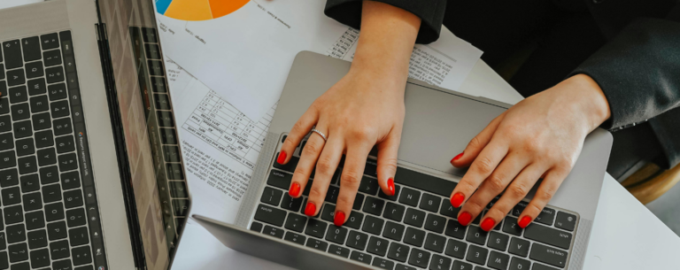 Person typing on a laptop with financial reports, spreadsheets, and a pie chart on the desk—suggesting business planning or financial analysis in a work or study setting.