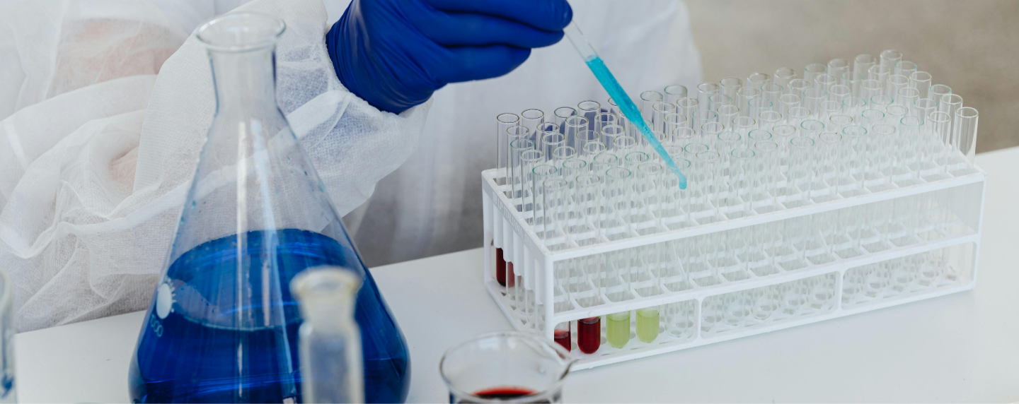 A scientist in protective clothing uses a pipette to transfer blue liquid into test tubes in a laboratory, with various glassware containing coloured solutions in the foreground.