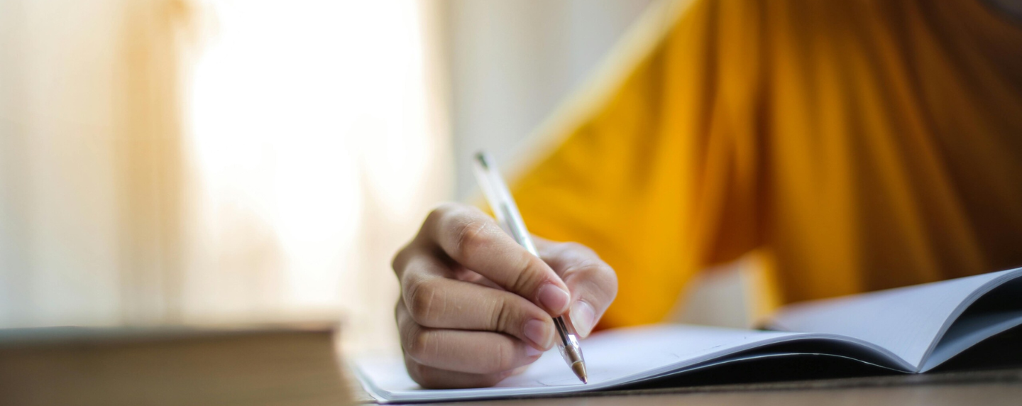 A student in a yellow shirt writing in a notebook at a desk, with a soft-focus book in the foreground and natural light coming through a window.