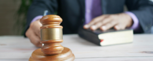 Close-up of a judge’s gavel on a wooden desk with a person in a suit holding a law book in the background, symbolising legal practice and justice.