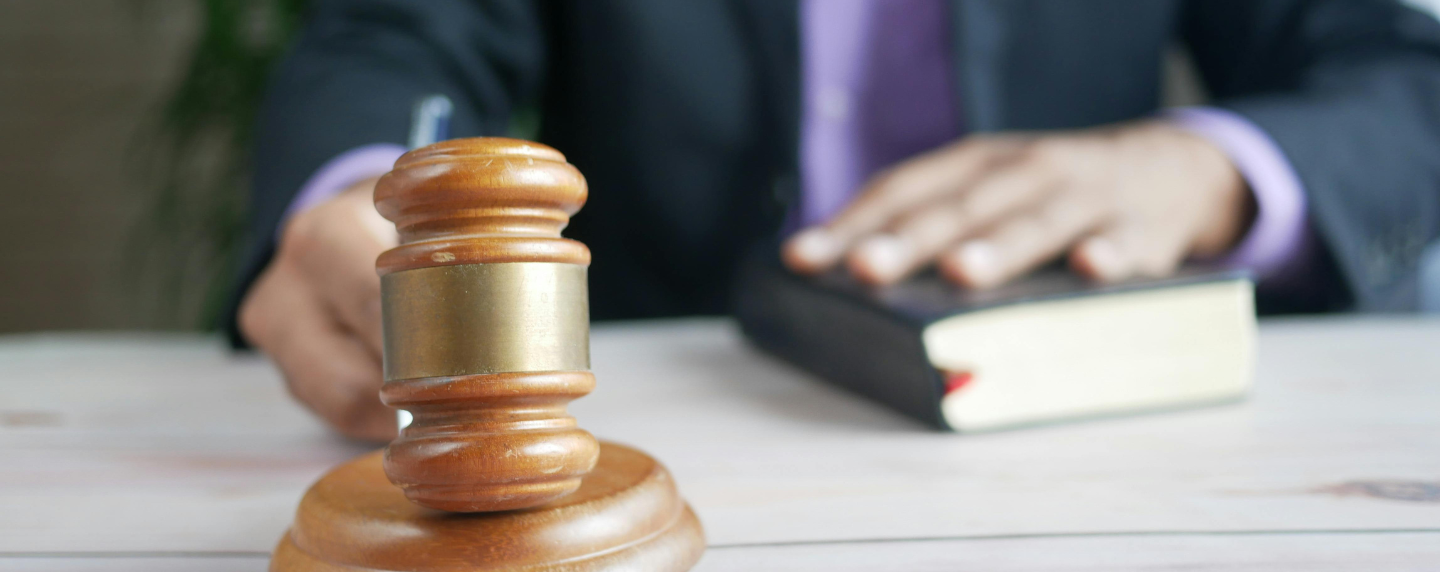 Close-up of a judge’s gavel on a wooden desk with a person in a suit holding a law book in the background, symbolising legal practice and justice.