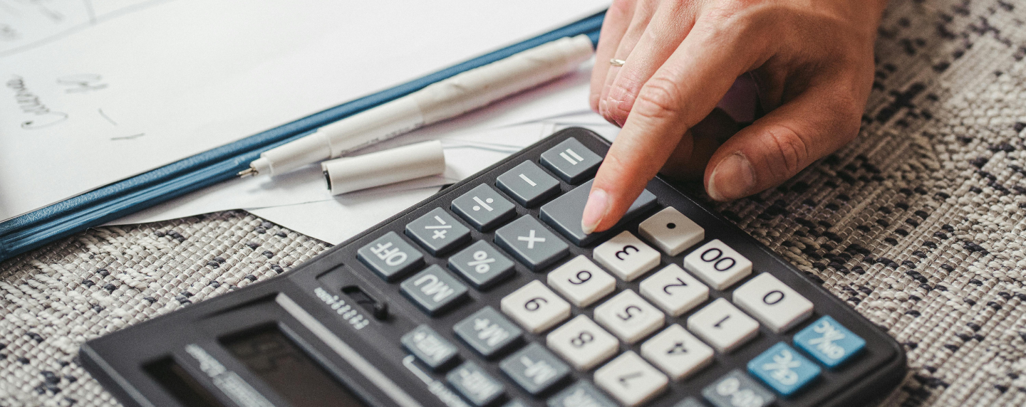 A close-up of a person using a calculator with worksheets and pens on a carpeted surface, representing focused mathematical problem-solving.
