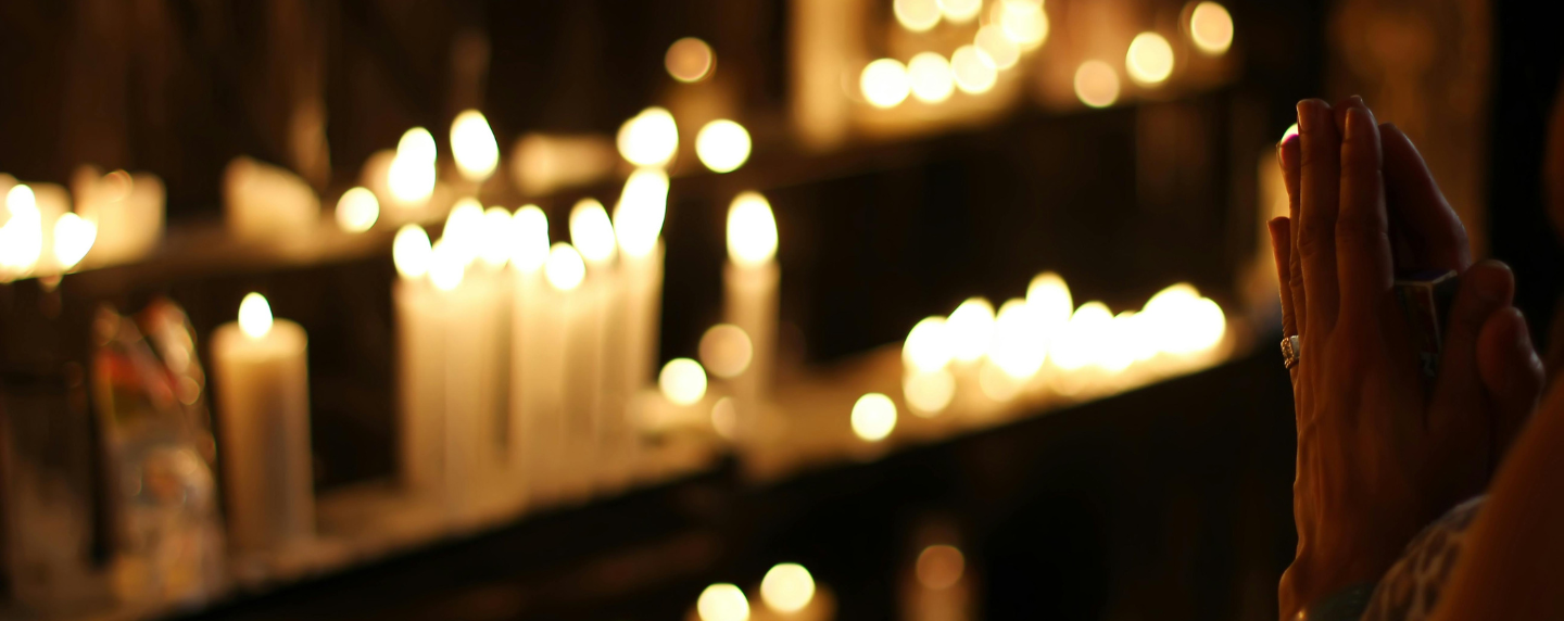 Close-up of hands held in prayer beside softly glowing candles in a dimly lit space, symbolising spirituality, reflection, and religious devotion.