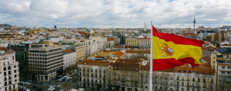 The Spanish flag waves above a cityscape in Madrid on a partly cloudy day, symbolising language, culture, and national identity