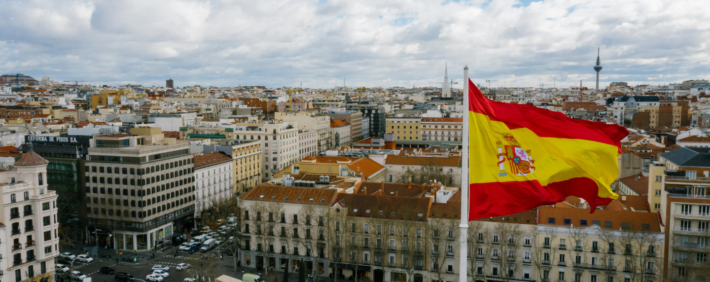 The Spanish flag waves above a cityscape in Madrid on a partly cloudy day, symbolising language, culture, and national identity