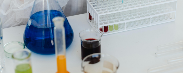 A close-up of a chemistry lab bench with colourful liquids in beakers, a conical flask filled with blue solution, and a rack of test tubes, representing chemical experiments and analysis.