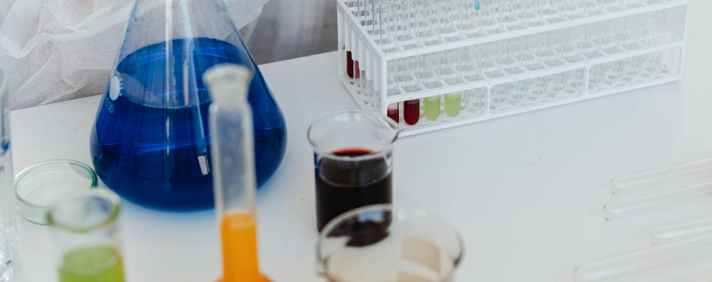 A close-up of a chemistry lab bench with colourful liquids in beakers, a conical flask filled with blue solution, and a rack of test tubes, representing chemical experiments and analysis.