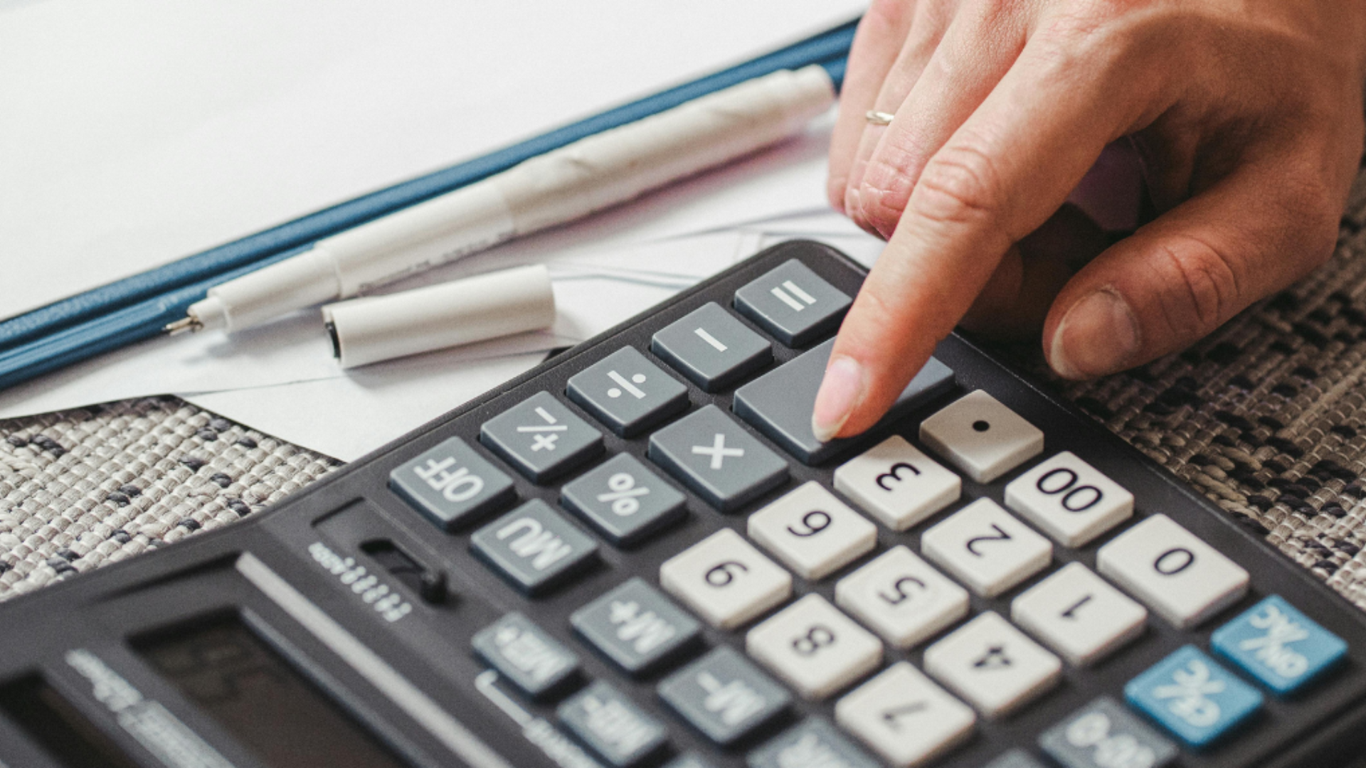 A close-up of a person using a calculator with worksheets and pens on a carpeted surface, representing focused mathematical problem-solving.