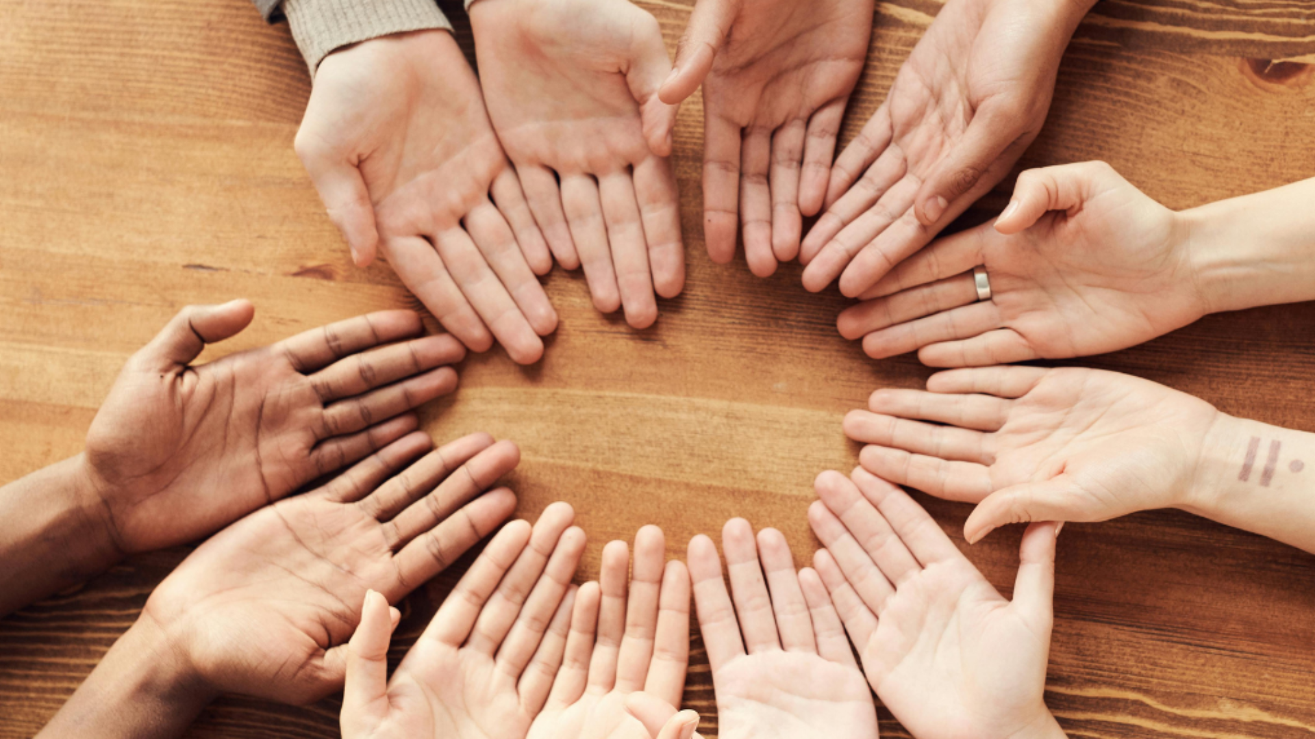 A group of diverse hands arranged in a circle on a wooden table, symbolising unity, inclusion, and social connection—key themes in sociology.