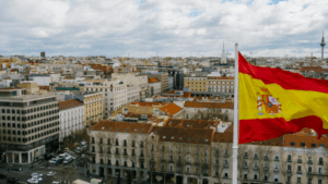 The Spanish flag waves above a cityscape in Madrid on a partly cloudy day, symbolising language, culture, and national identity