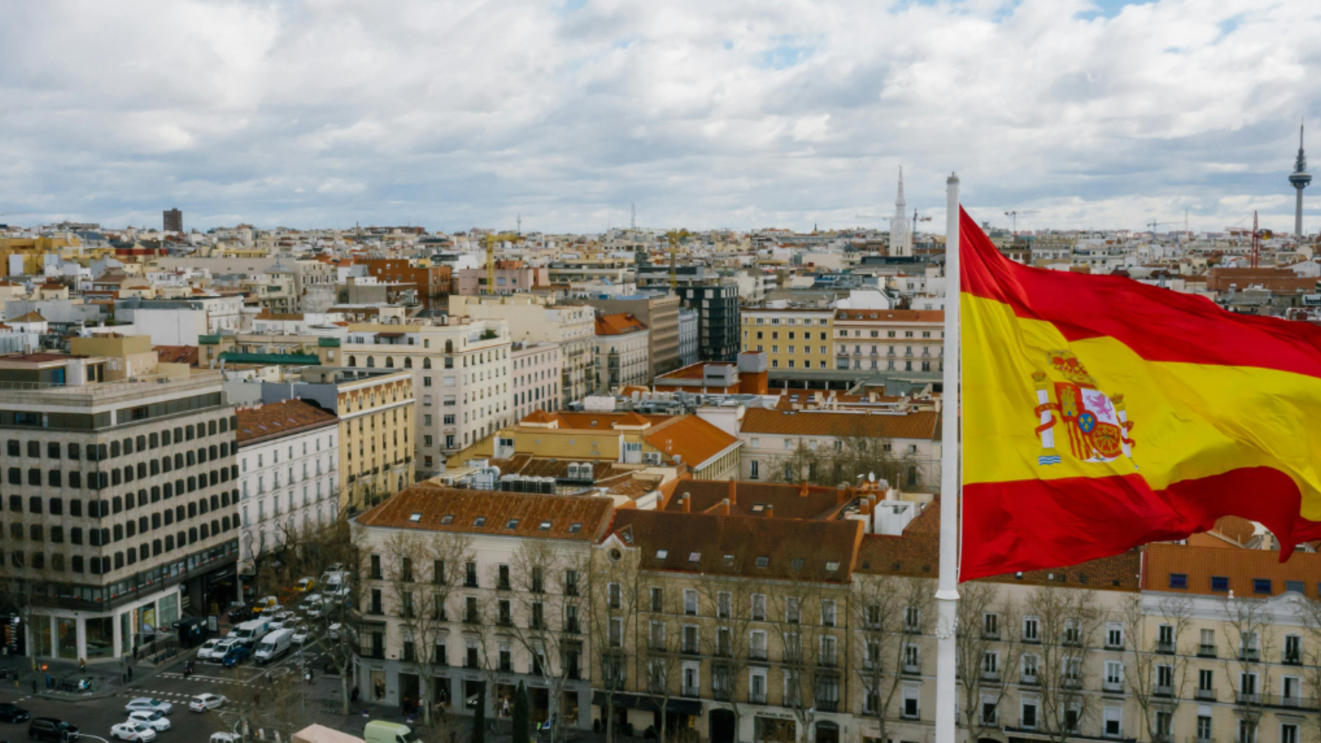 The Spanish flag waves above a cityscape in Madrid on a partly cloudy day, symbolising language, culture, and national identity