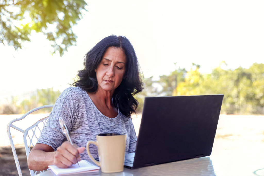 Older learner sits outside with her laptop, writing down notes in a notebook.