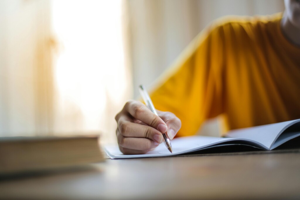 A student in a yellow shirt writing in a notebook at a desk, with a soft-focus book in the foreground and natural light coming through a window.