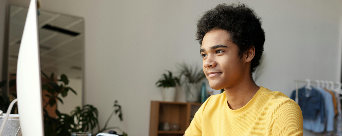 A young student wearing a yellow top smiles while working at a computer in a relaxed home study space, representing the flexibility and independence of online A level learning.