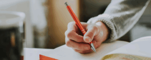 A hand writing in a notebook with a red pen, symbolising the study of language, grammar, and communication.