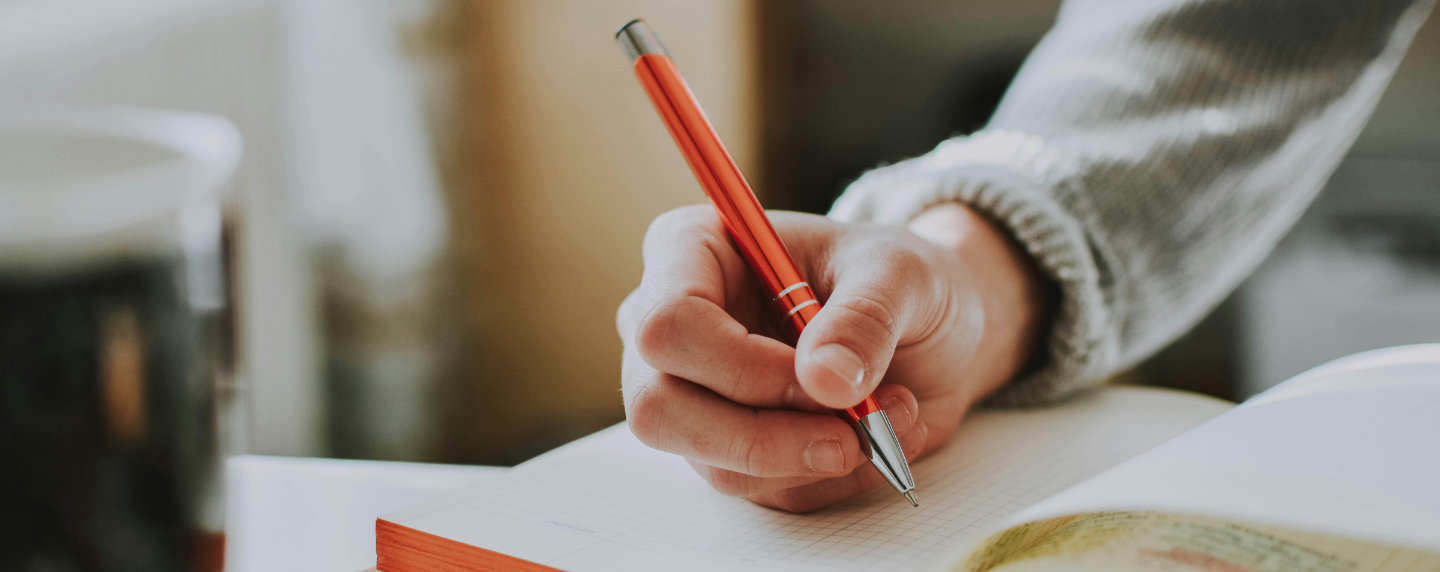 A hand writing in a notebook with a red pen, symbolising the study of language, grammar, and communication.