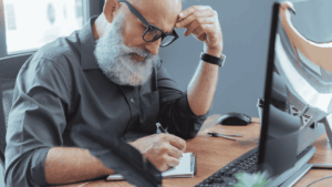 An older man with grey hair and glasses sitting at a desk, focused as he writes in a notebook beside a desktop computer—symbolising lifelong learning and academic determination.