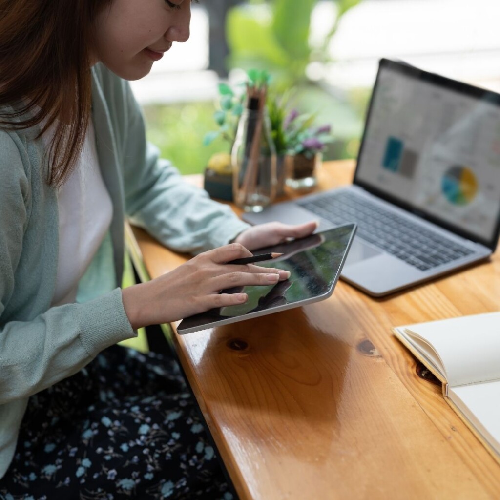 Business management overview, online courses with NEC. Female working on a tablet and laptop sat on the desk.