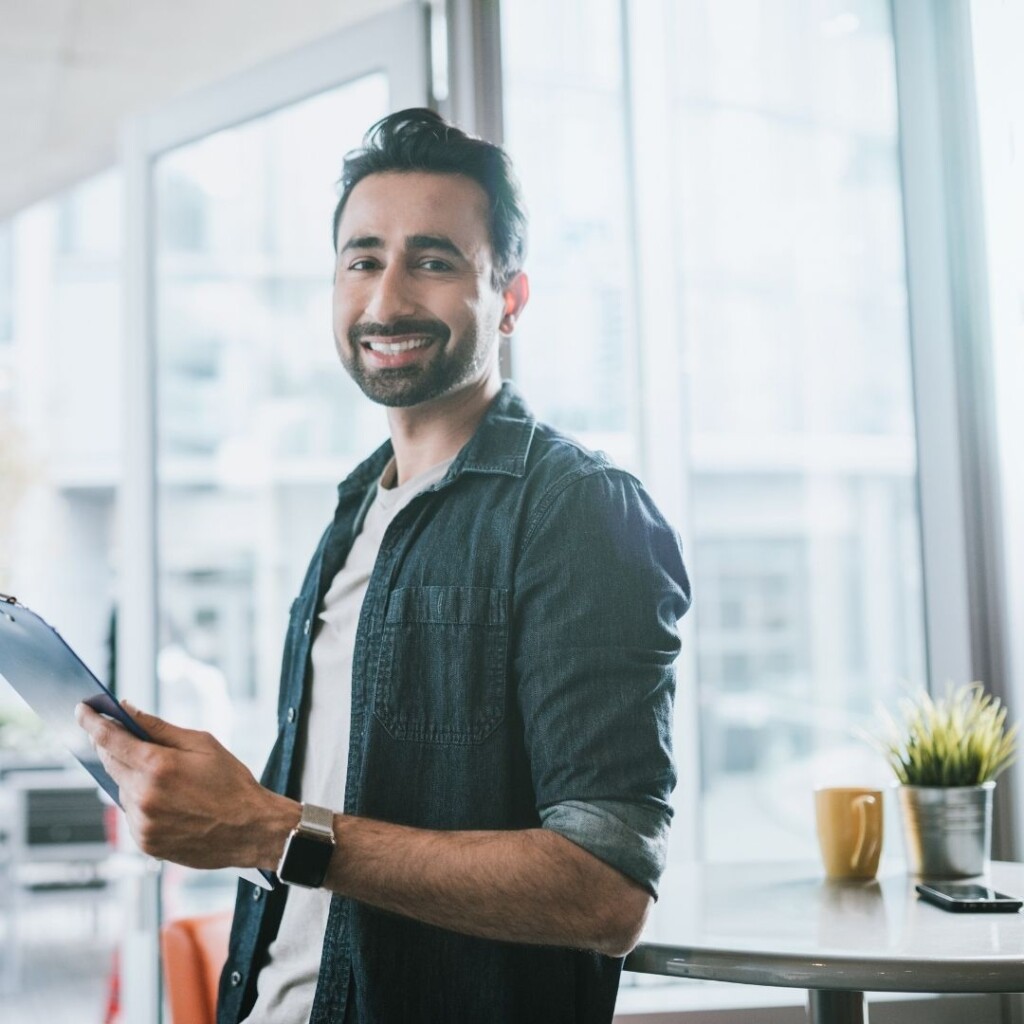 NEC experts in online learning. Man stood with clip board, stood in front of a desk in front of a large window