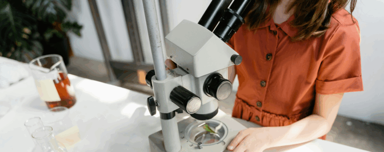 Student in a rust-red dress using a microscope to examine a leaf in a science lab setting, surrounded by beakers and scientific equipment.