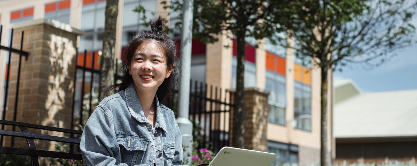 Young student smiling outdoors while working on a laptop, symbolising the flexibility and independence of NEC’s online Fast Track A level programme.