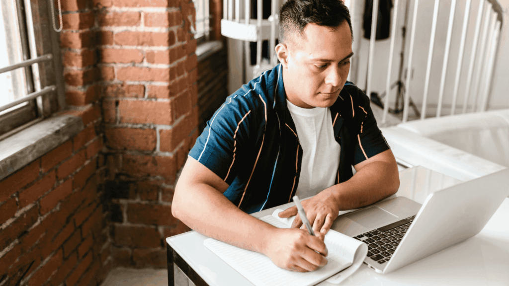 A focused adult student sitting at a desk in a bright room, writing notes in a notebook while looking at an open laptop—symbolising flexible learning and mature study.